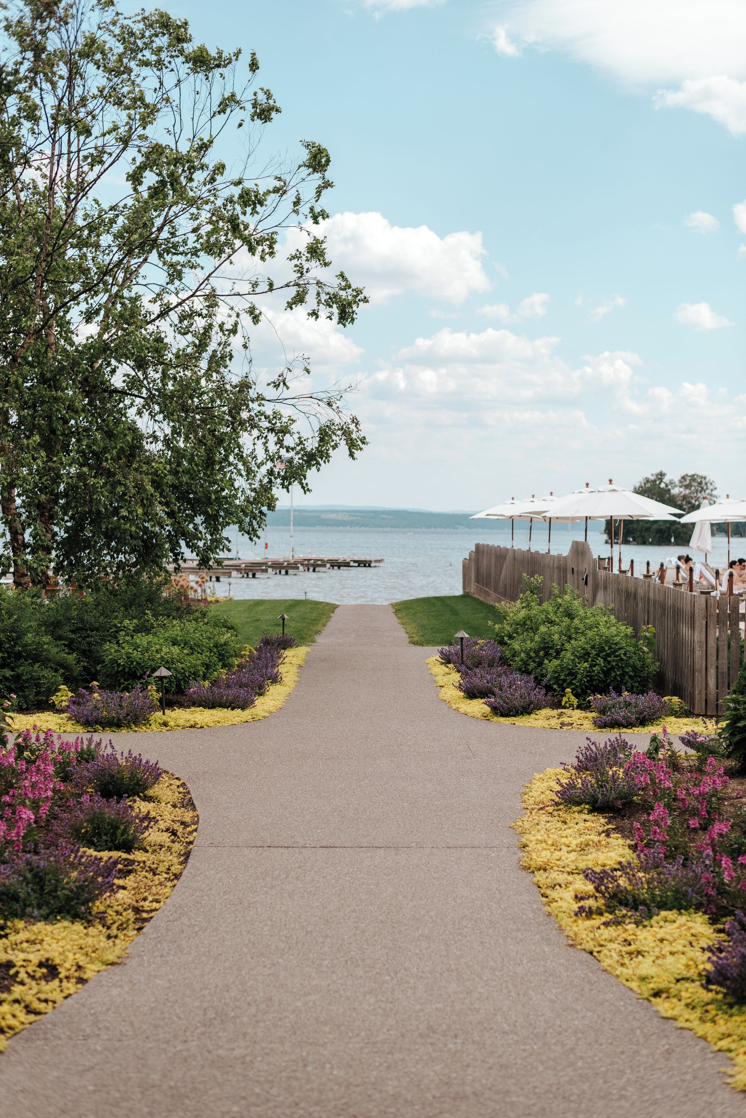A paved pathway leading to a waterfront area with a wooden fence and umbrellas.