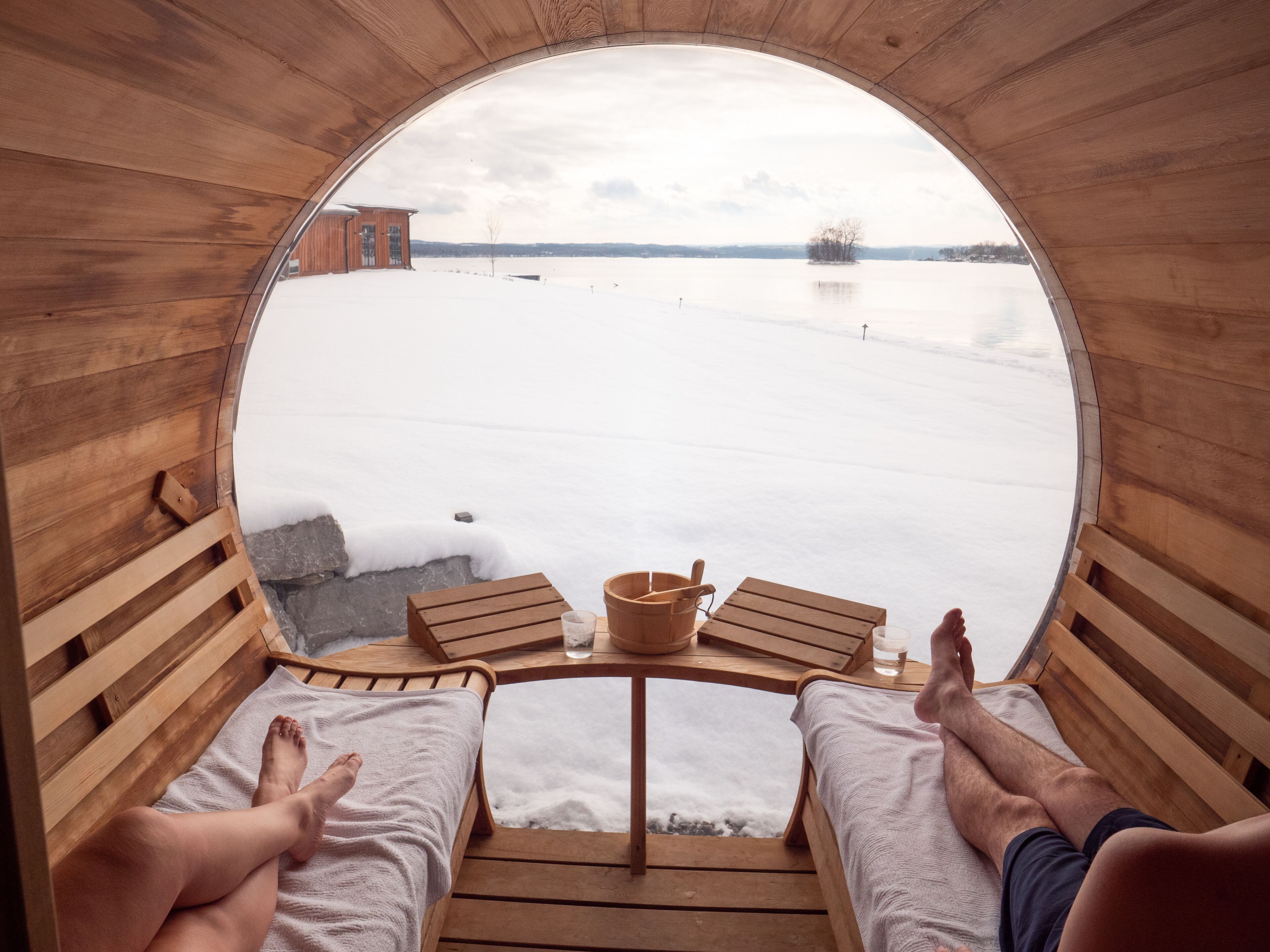 Una cabaña de madera con un ventanal circular que da a un paisaje nevado. En el interior, hay dos bancos de madera con cojines y una pequeña mesa con un balde y un vaso.