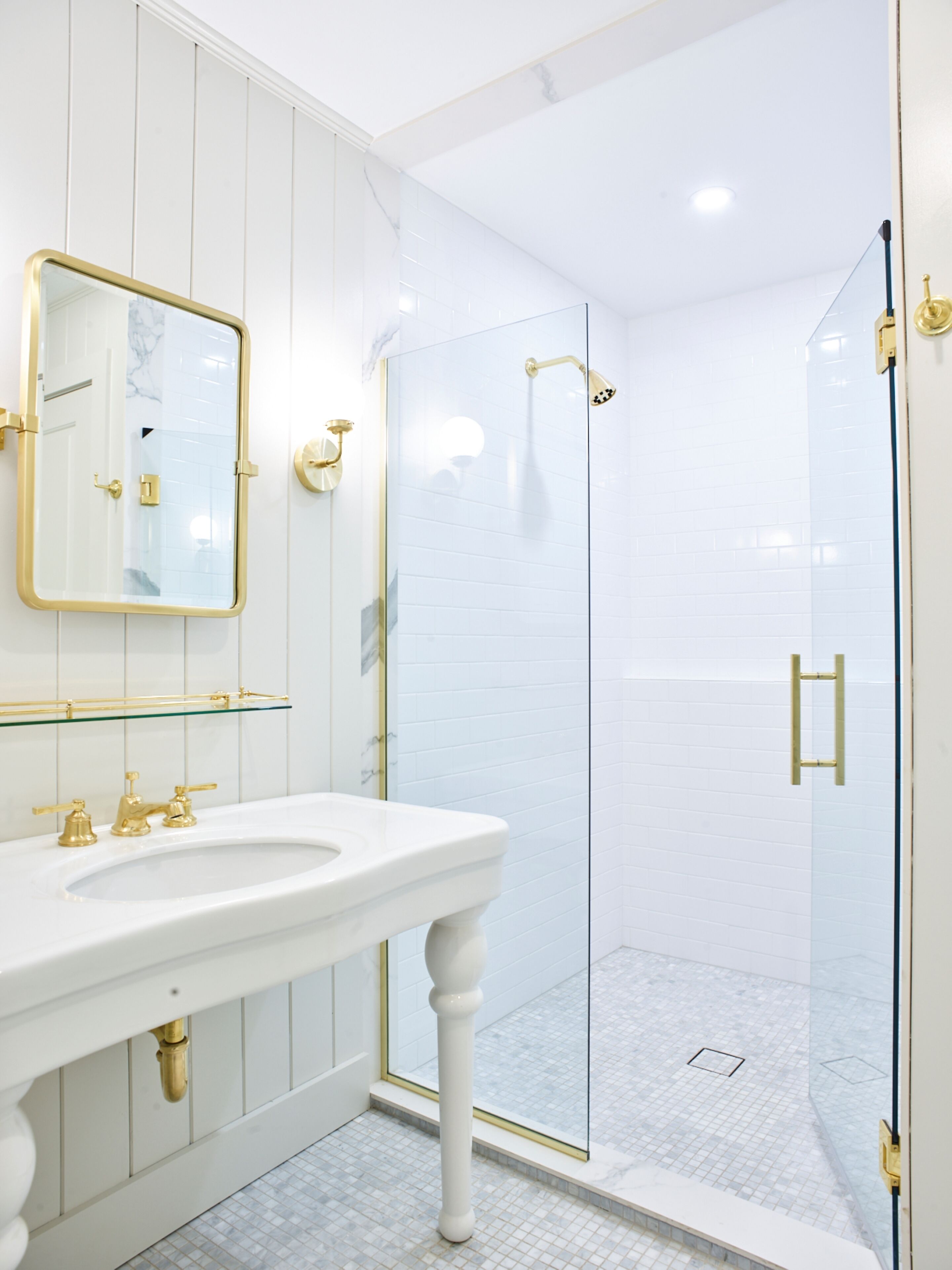 Une salle de bain moderne avec un lavabo blanc, des robinetteries dorées, une douche en verre et un sol carrelé en marbre.
