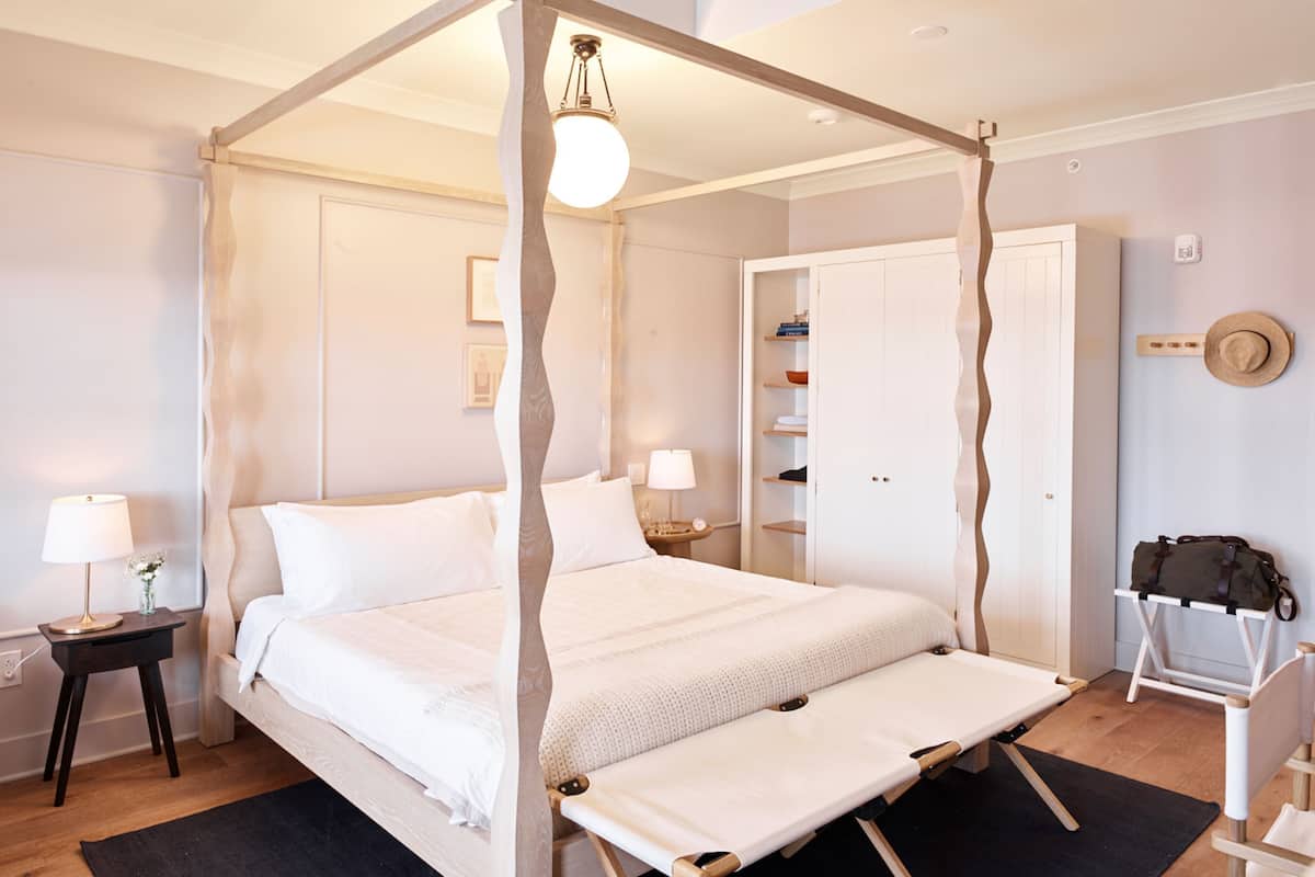 Ein Himmelsbett mit Baldachin, Nachttische mit Lampen, ein Nachttischregal, ein Kleiderschrank und ein Koffer auf einem Stuhl.
