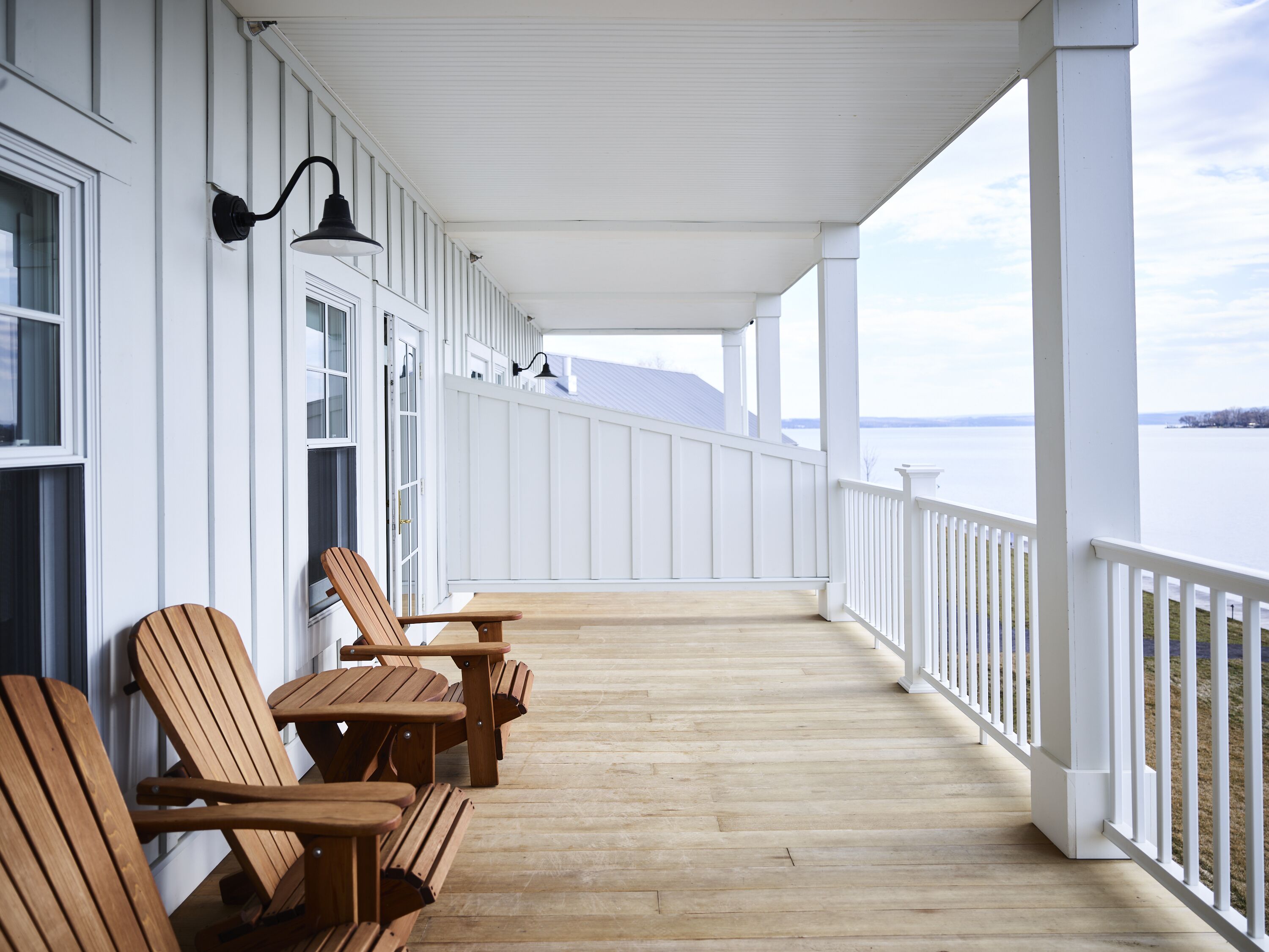 A covered porch with wooden chairs and tables, white railings, and a view of a body of water.