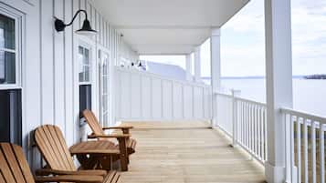 A covered porch with wooden chairs and tables, white railings, and a view of a body of water.