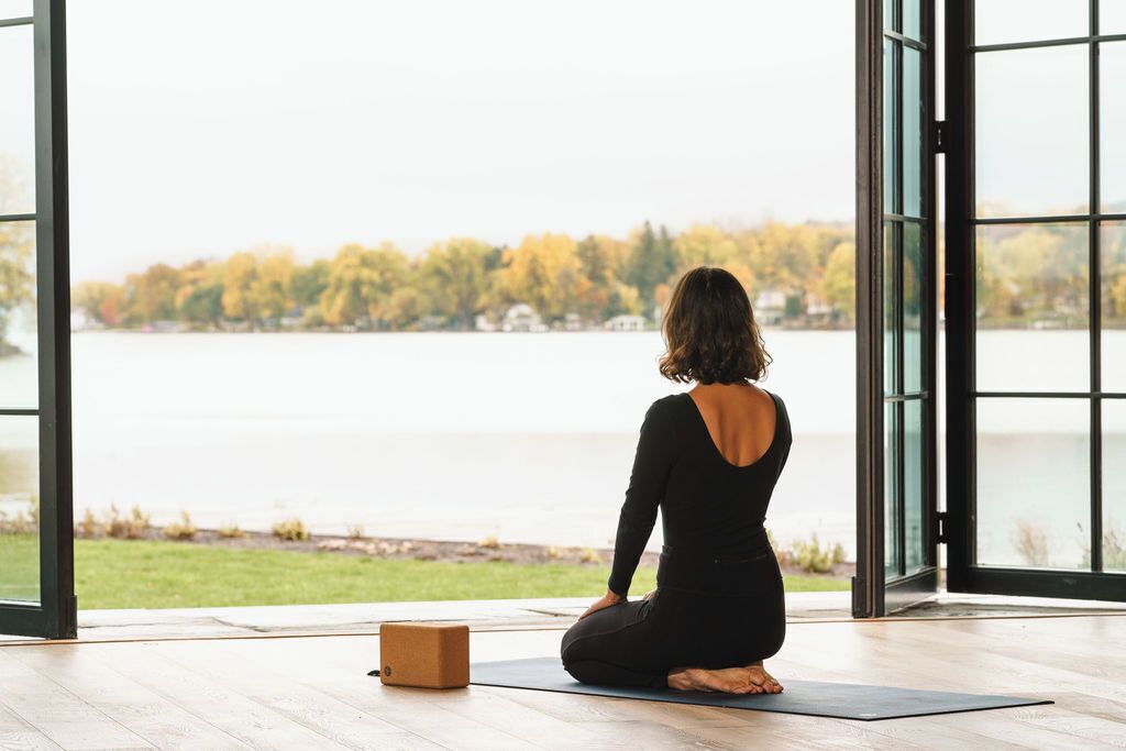 Eine Person sitzt auf einer Yogamatte in einem Raum mit großen Fenstern mit Blick auf einen See und Bäume.