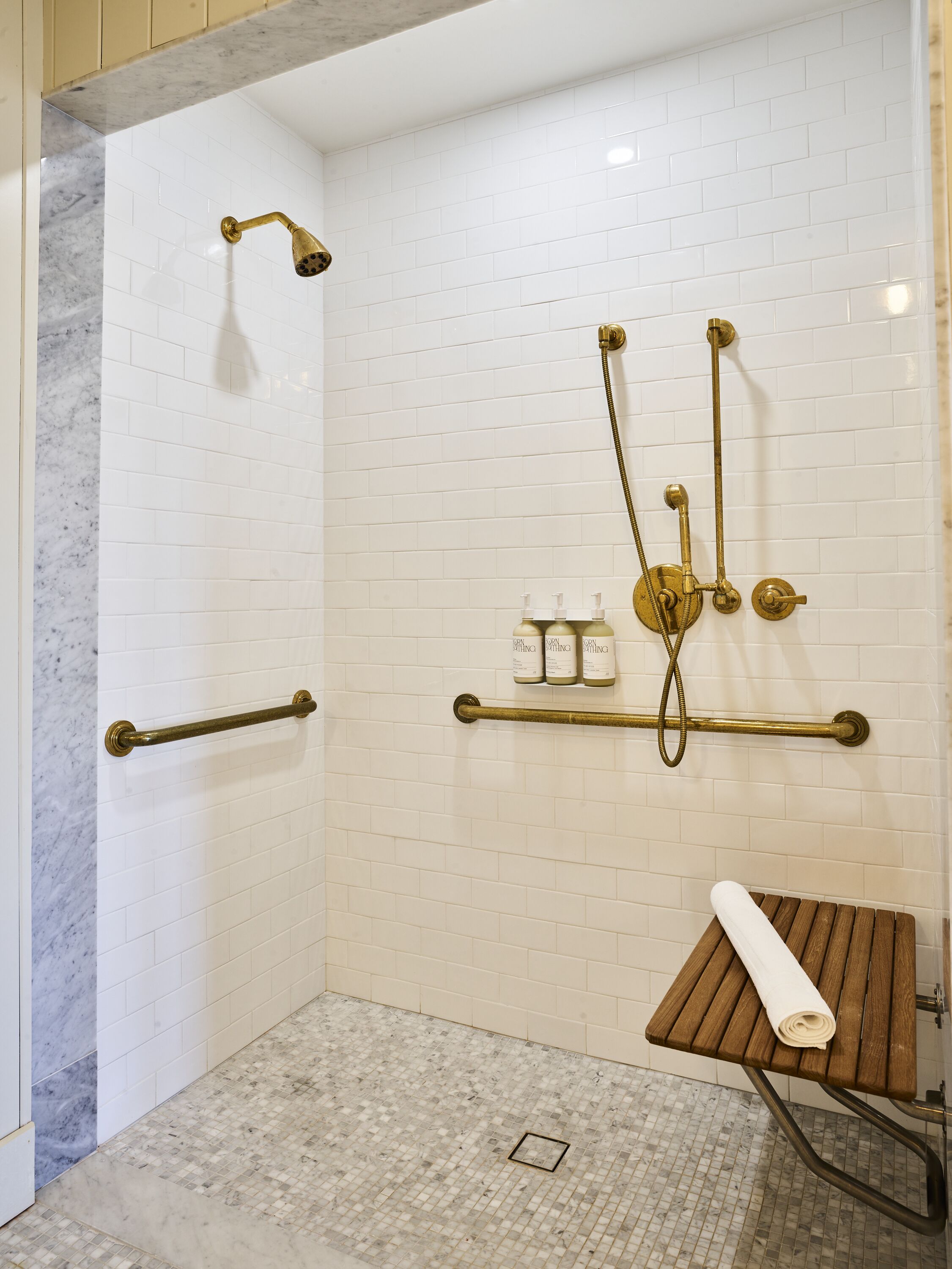 Une douche à l’italienne avec des murs recouverts de carreaux blancs, un sol en marbre et un banc en bois.