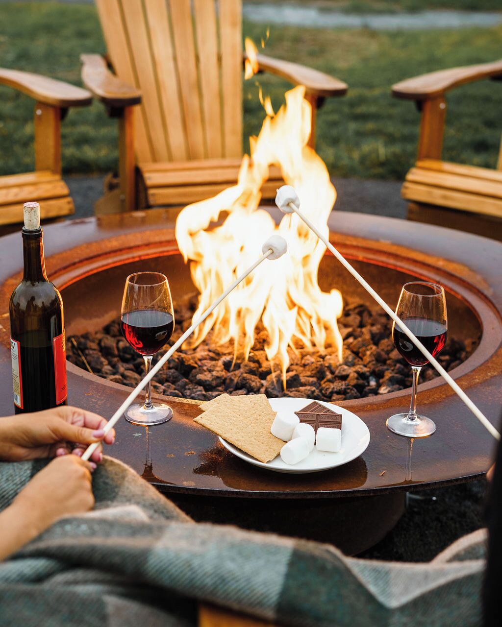A fire pit with two glasses of red wine, marshmallows, and a chocolate bar.