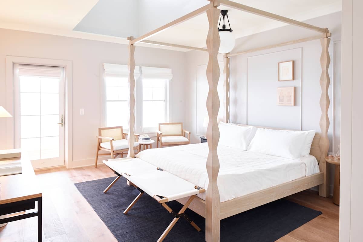 Ein Bett mit Baldachin, dessen Gestell aus Holz besteht, mit weißer Bettwäsche, zwei Holzstühle, ein kleiner Tisch und ein schwarzer Teppich in einem hellen Raum mit weißen Wänden und einer Tür.