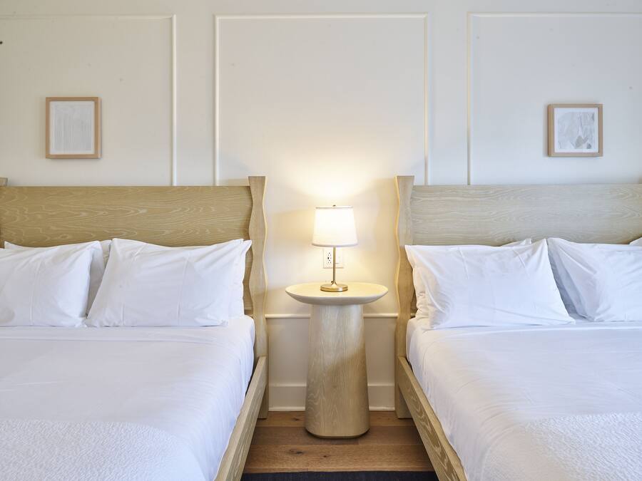 A hotel room with two beds, a round bedside table with a lamp, and two framed artworks on the walls.