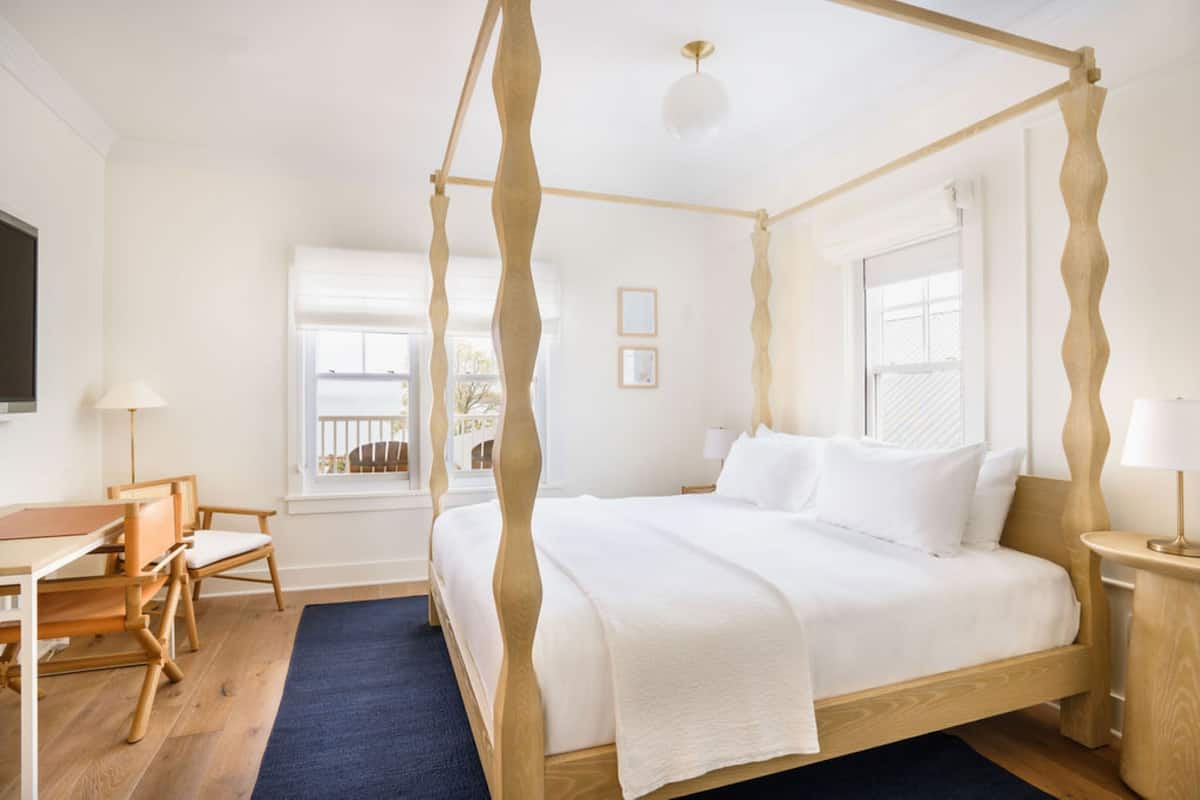 Ein Bett mit Baldachin, dessen Gestell aus Holz ist, ein Nachttisch mit Lampe, ein Sessel und ein Schreibtisch ebenfalls mit Lampe.