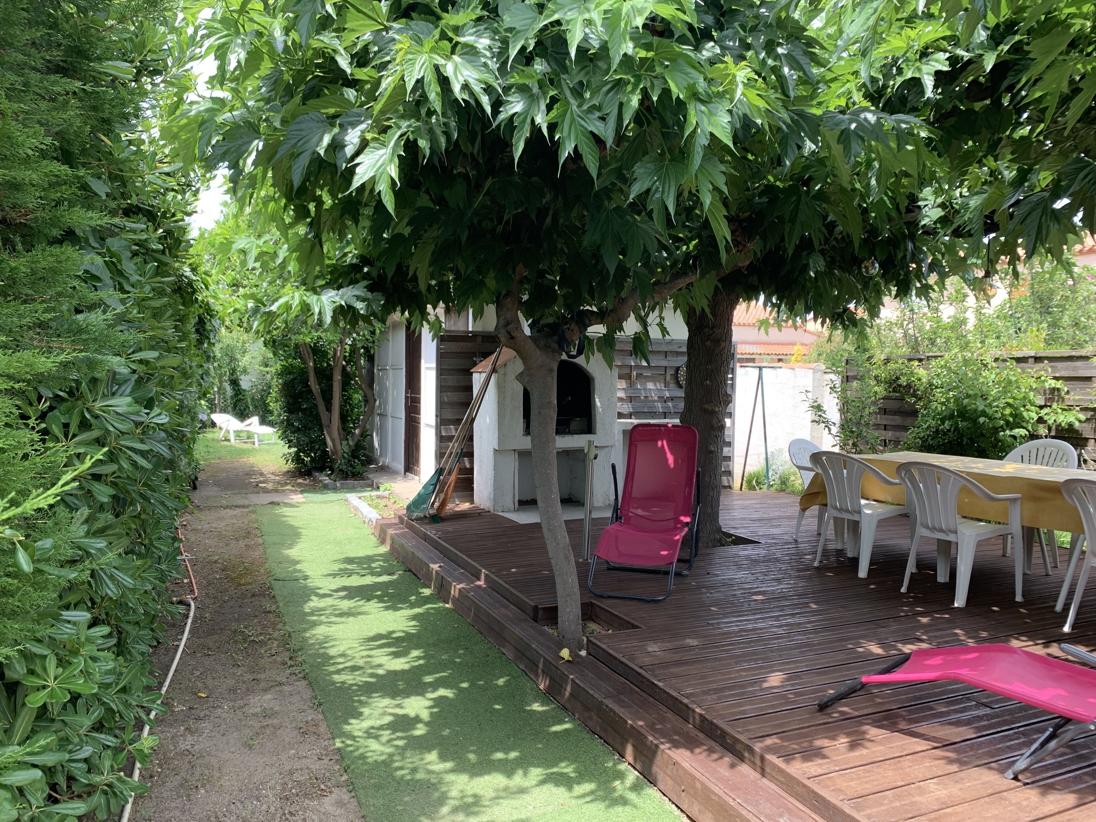 En træterrasse med et bord og stole, omgivet af frodigt grønt og et træ.