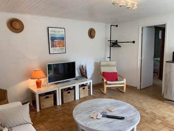 Een woonkamer met een televisie, een ronde salontafel, rieten manden, een stoel met een rode kussen en een hoed aan de muur.