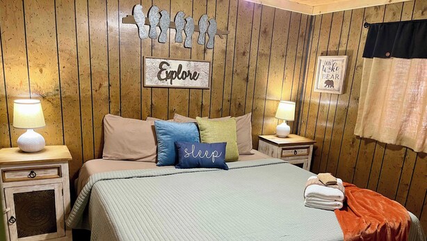 A bedroom with a bed, two bedside tables, a wall with wooden paneling, and a sign that says 'Explore'. - Pet-Friendly Sleepin Inn Cabin w/ Hot Tub & Fireplace (Ruidoso)