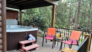 A wooden deck with a hot tub, two chairs, a table with drinks, and a wooden bench. - Pet-Friendly Sleepin Inn Cabin w/ Hot Tub & Fireplace (Ruidoso)