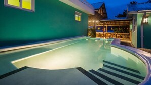 Ein Schwimmbecken mit glatter Wasseroberfläche, umgeben von einem Gebäude mit grüner Fassade und einer überdachten Sitzterrasse.