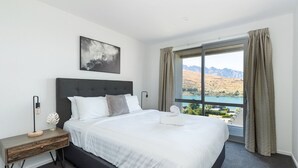 Habitación de hotel con una cama grande, una mesita de noche, una lámpara, un cuadro y vistas a las montañas y al agua.