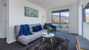 Una sala de estar moderna con un sofá gris, un sillón azul, una mesa de centro y vistas a montañas y agua a través de ventanas amplias.