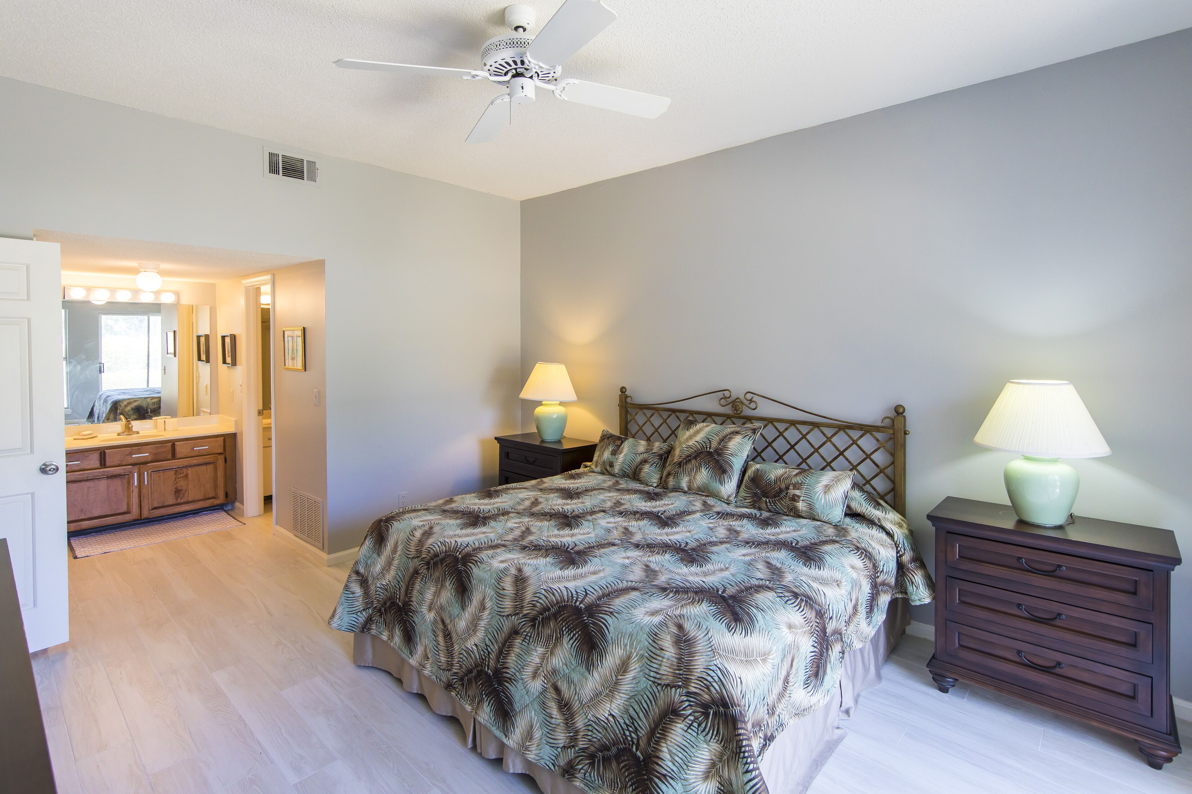 Une chambre à coucher avec un lit, deux tables de chevet avec des lampes, un ventilateur de plafond et une salle de bain visible à travers une porte ouverte.