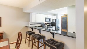 A modern kitchen with white cabinets, a black countertop, and a stainless steel refrigerator. There are three bar stools and a wooden dining table with chairs. - Deeded Beach Access, Community Pool, Screen Porch, Walk to Shops and Restaurants (Santa Rosa Beach)