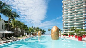 Piscina com espreguiçadeiras e palmeiras, cercada por um prédio alto de hotel.