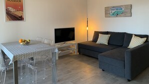 A modern living room with a grey sofa, a TV, a wicker pendant light, a table with a fruit bowl, and a wall with a poster and a car painting. - Apartment T3 with terrace, 100 m from the beach and 5 min by bike from the lake (Soorts-Hossegor)