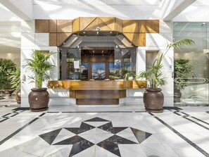 Uma área de recepção com design moderno, com balcão de atendimento, grandes plantas em vasos e piso de azulejos geométricos.