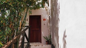Una escalera estrecha que lleva a una puerta de madera, con una planta en maceta a la derecha y plantas de bambú a la izquierda.