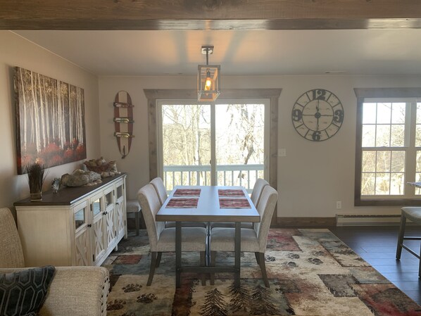 A dining area with a table, chairs, a clock, and a view of trees through large windows.