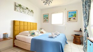 Un dormitorio con cabecera amarilla, colcha azul y obras de arte con motivos tropicales en las paredes.