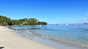 A beach with clear water, boats, and a house on a hill.