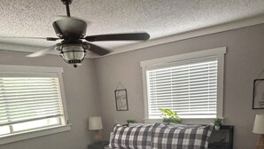 Un dormitorio con cama, ventilador de techo, dos ventanas con persianas, mesita de noche con lámpara y un pequeño banco.