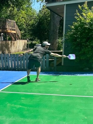 A person playing tennis on an outdoor court with a wooden fence and trees in the background.