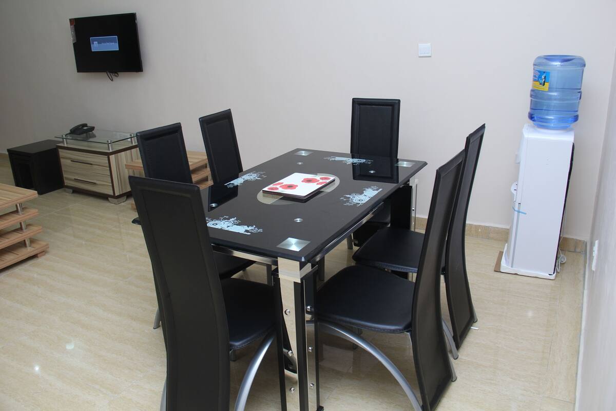 A conference room with a glass table, black chairs, a white cabinet with a water dispenser, and a mounted TV.