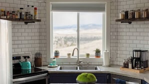 Uma cozinha com azulejos brancos tipo metrô, uma janela com vista para uma paisagem montanhosa, uma pia e diversos utensílios e eletrodomésticos de cozinha.