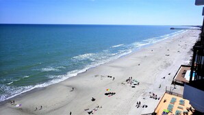 A beach with a pier extending into the ocean, a sandy shore with people and beach chairs, and a building with balconies overlooking the beach. - Stunning Royal Garden Unit 1002 (Murrells Inlet)