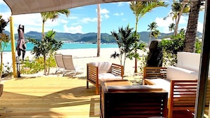 Eine Holzterrasse mit Gartenmöbeln, darunter eine Couch und Stühle, mit Blick auf einen Strand, im Hintergrund Palmen und Berge.