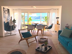 Un salón con sofá, mesa de centro y comedor con mesa y sillas. Se ve la playa y las montañas a través de amplios ventanales.