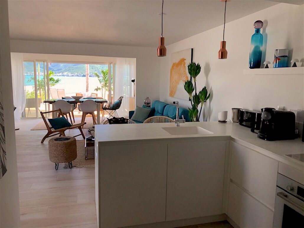 A modern kitchen with a central island, a dining area with chairs and a table, and a view of the beach through large windows.