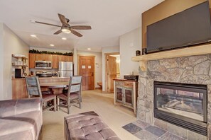 A modern kitchen with wooden cabinets, a dining area with a wooden table and chairs, a fireplace, and a flat-screen TV mounted above it.