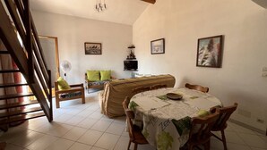 Un comedor con una mesa cubierta con mantel, un sofá, un televisor y cuadros en la pared.