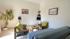 Una habitación pequeña y bien iluminada con una cama, dos sillas, una mesa, un ventilador, un televisor y una planta.