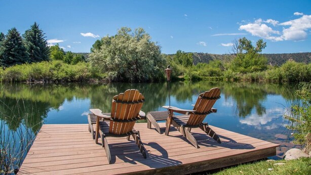 A wooden dock with two Adirondack chairs facing a calm lake, surrounded by lush greenery and trees.