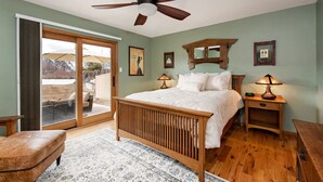 A bedroom with a wooden bed, a ceiling fan, a nightstand with a lamp, and a large window with a view of the outdoors.