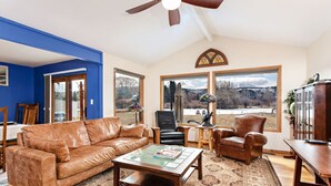 A living room with a brown leather sofa, a coffee table, and a patterned rug. There are two chairs, a lamp, and a view of a snowy landscape through large windows.