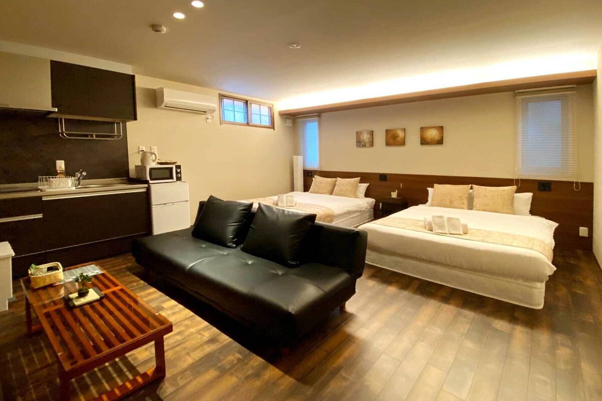 A modern hotel room with a black leather sofa, a wooden coffee table, two beds with white bedding, a kitchenette, and a small dining area.