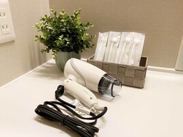 Ein Haartrockner, ein Mehrfachsteckdosenadapter, ein Topfpflanze und eine Aufbewahrungsbox mit Gegenständen auf einer weißen Arbeitsplatte.