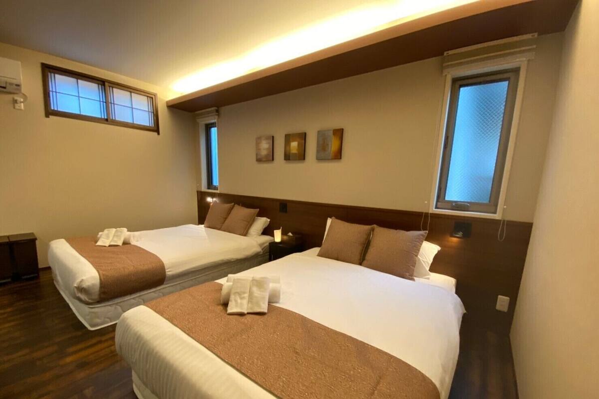 A hotel room with two beds, each with a brown blanket and white linens. There are bedside tables with lamps, a window with blinds, and artwork on the wall.