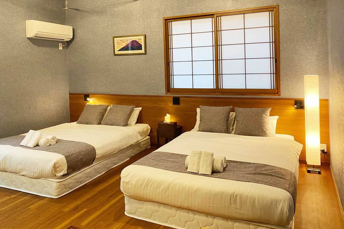 A hotel room with two beds, a large window, a wall-mounted air conditioner, and a bedside table with a lamp.