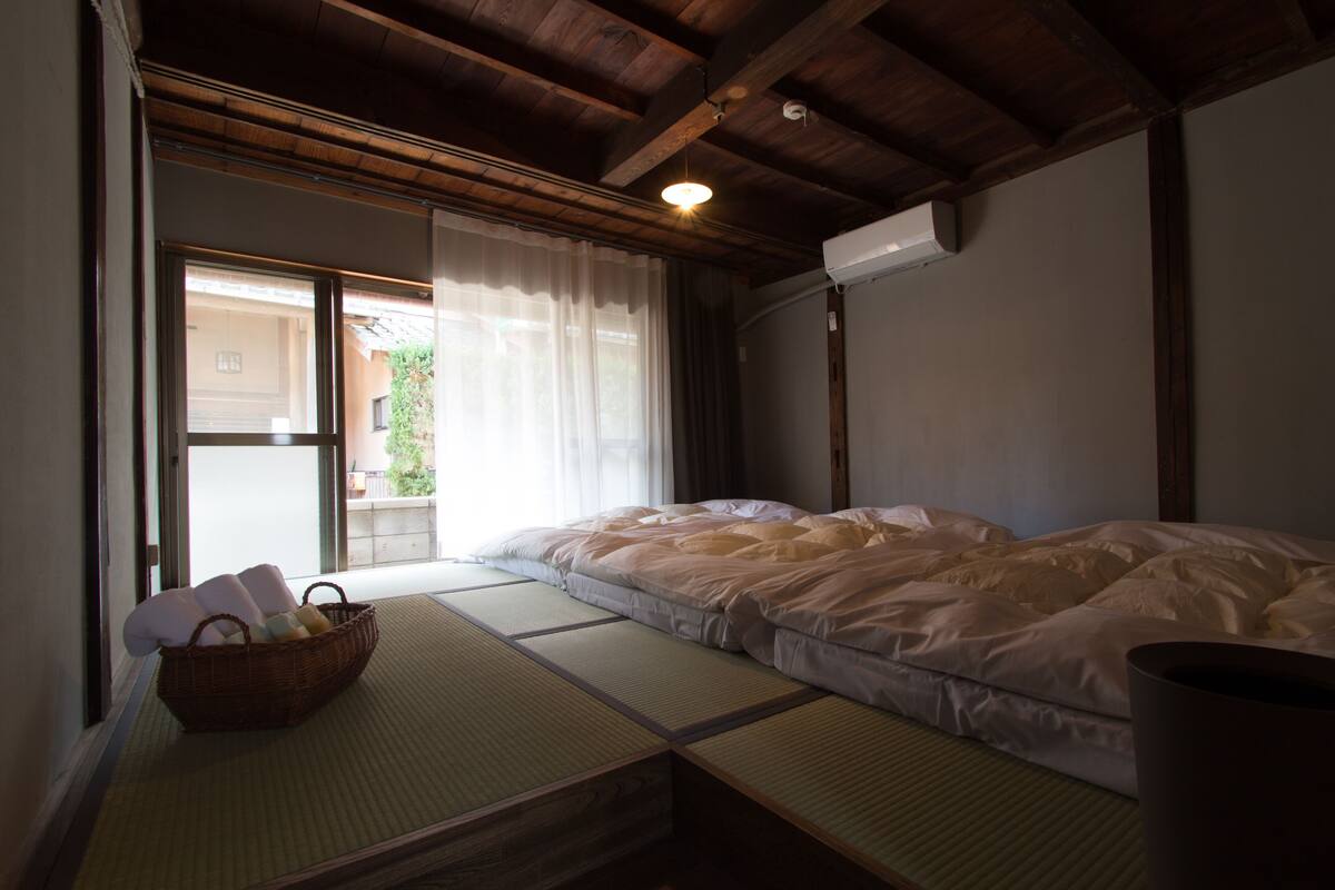 A traditional Japanese room with tatami flooring, a bed with white bedding, a wooden ceiling, and a sliding door leading to an outdoor area.