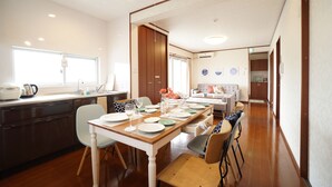 A dining area with a wooden table set for six, surrounded by chairs. A kitchen area with a window and a sink is visible to the left. - PINK Suite HOUSE (Nanjo)