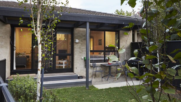 A modern house with a covered patio, a dining area, and a visible interior with a sofa and a table. - Brantome (Bright)