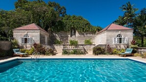 A swimming pool with two stone buildings, each with a red roof and arched windows, surrounded by greenery. - Porters Villa by Blue Sky Luxury (Holetown)